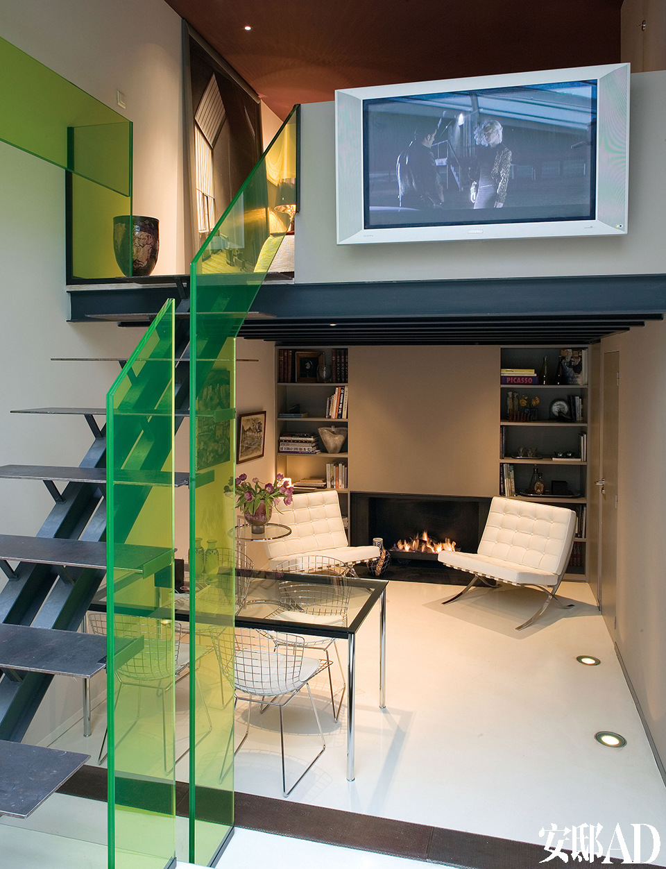 金属楼梯的每一级台阶都由钢条铺设，绿色玻璃扶手成为整个家的点睛之笔。两把白色巴塞罗那扶手椅与壁炉搭配成一个温暖的阅读区。从楼梯拾级而上，就是夹层卧室。夹层上的镀锌的陶罐来自Domani。